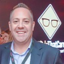 Smiling man in a blazer at a conference, with a diamond-shaped logo on a screen behind him.