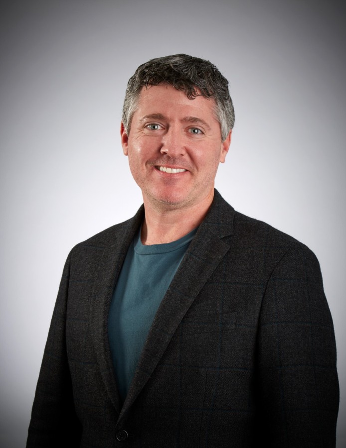 Smiling man in a dark blazer and teal shirt, posing for a professional headshot against a neutral gradient background.