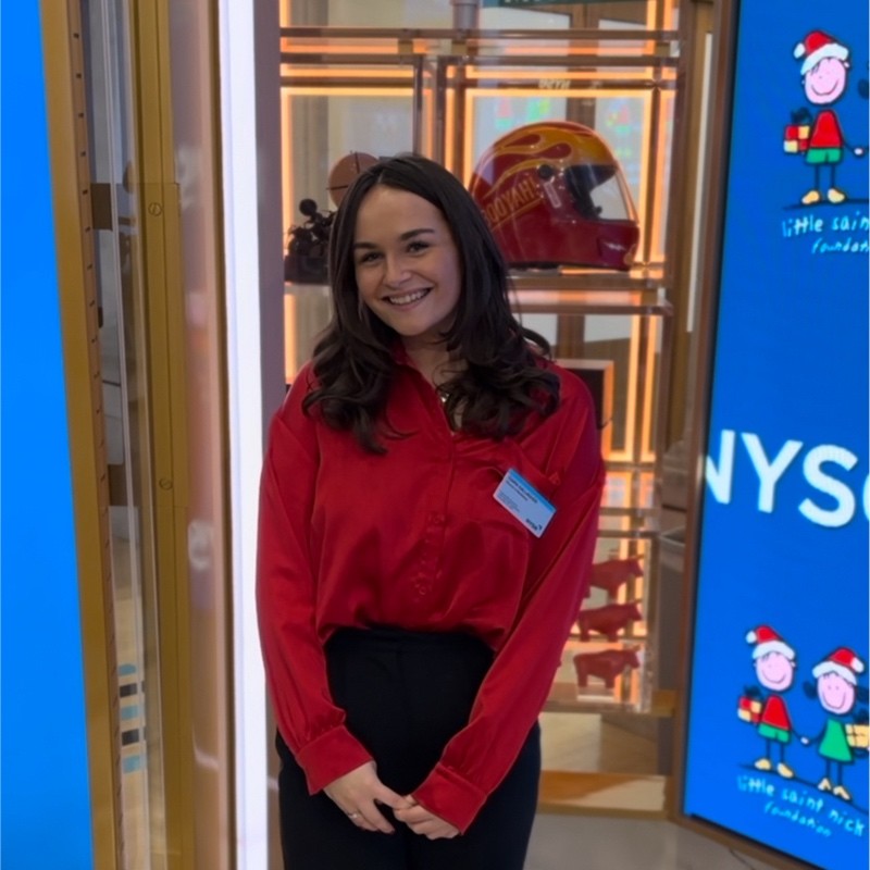 Smiling woman in a red blouse with a name badge stands in a bright showroom with helmet displays behind her.