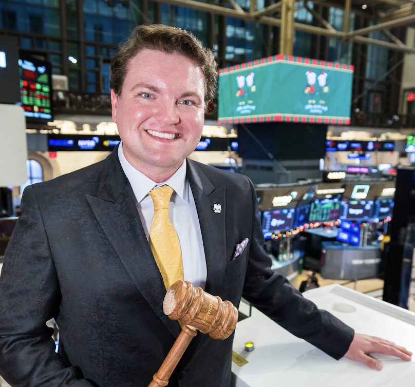 Smiling man in a dark suit and yellow tie standing on a trading floor, holding a wooden gavel.