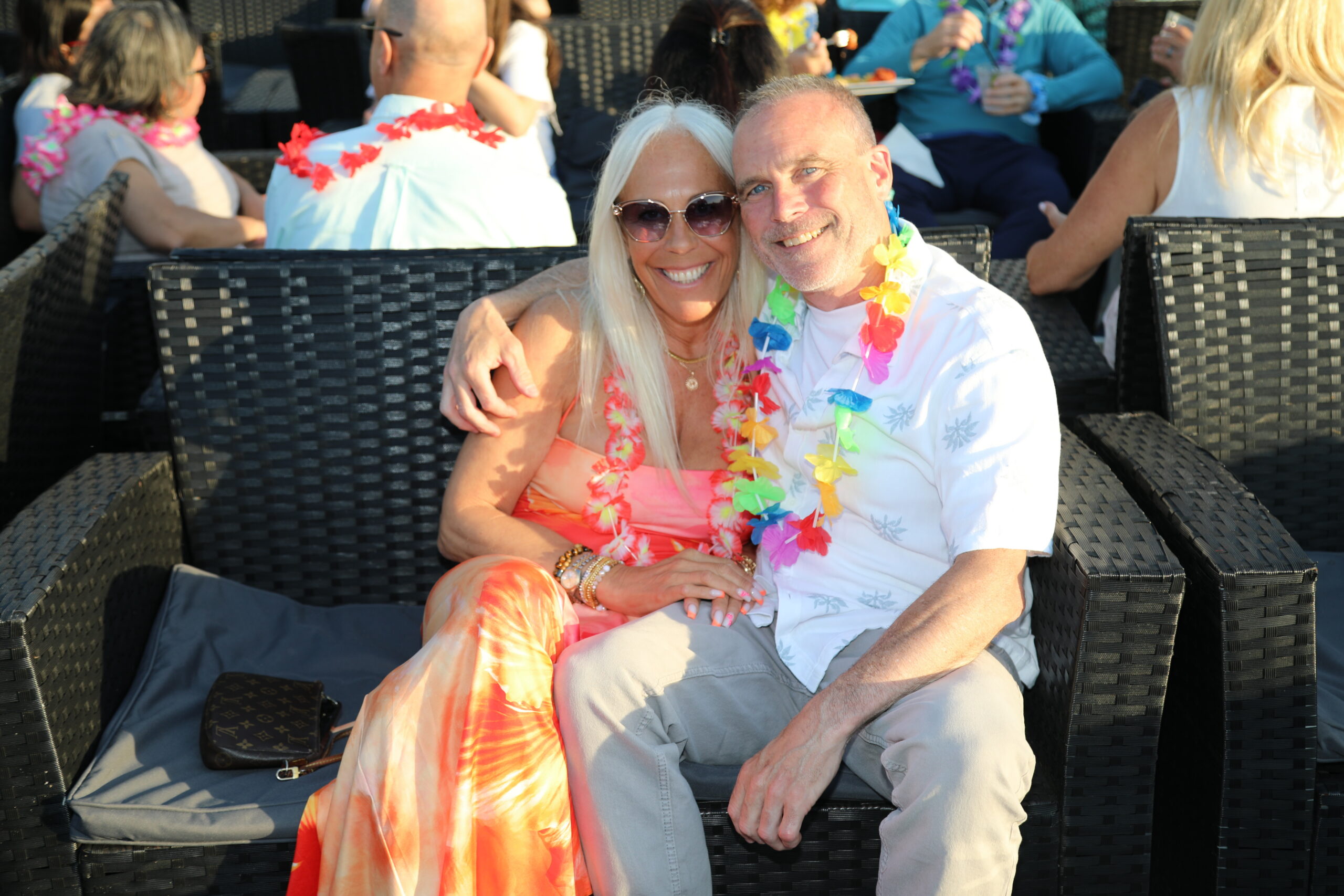 Couple sitting close together at a tropical party, wearing leis and bright clothing, smiling at the camera.