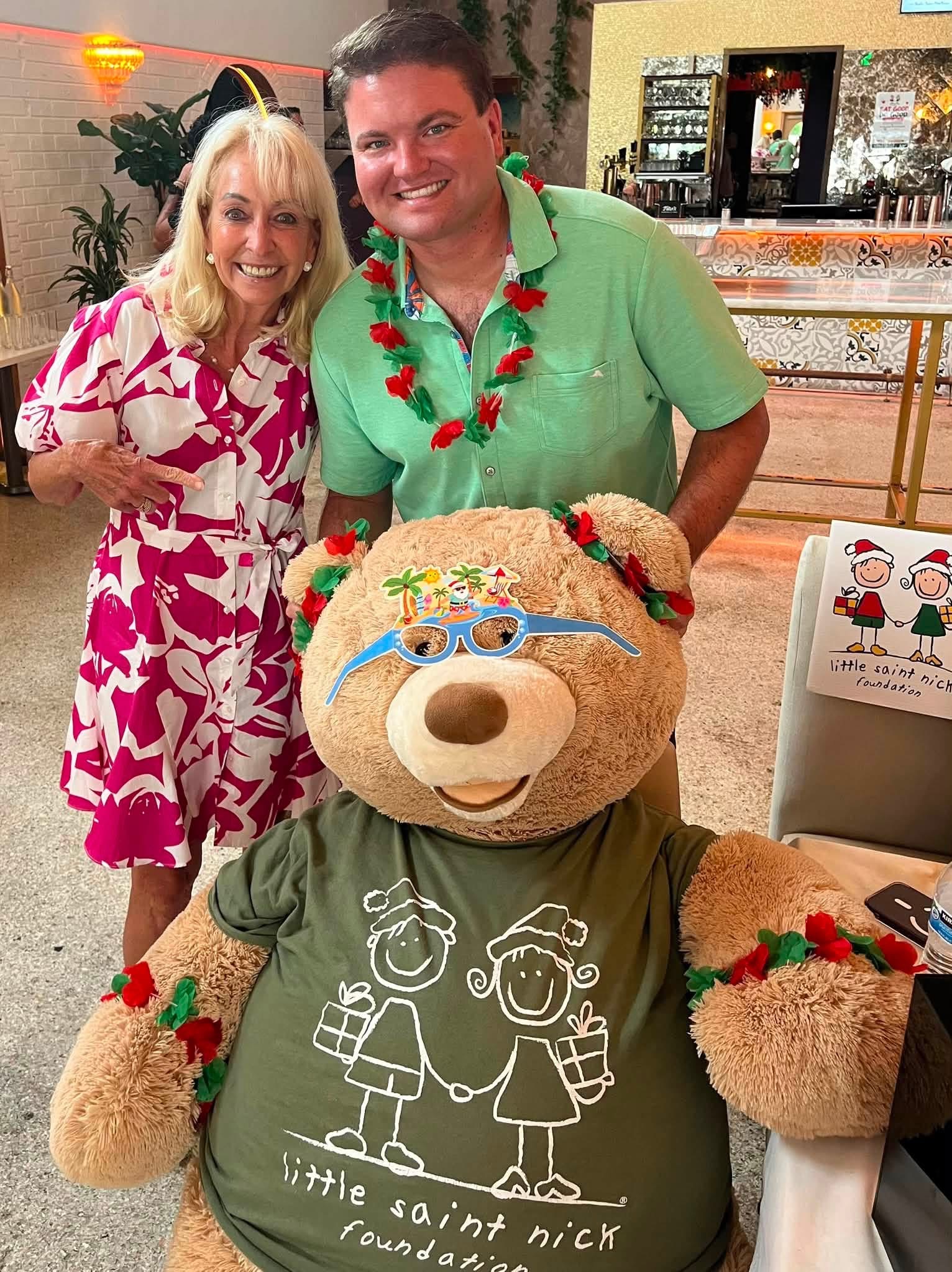 Smiling woman in a pink and white dress posing with a man wearing a green shirt and a tropical lei, beside a large teddy bear with a gift-themed illustration on its shirt at a festive venue.