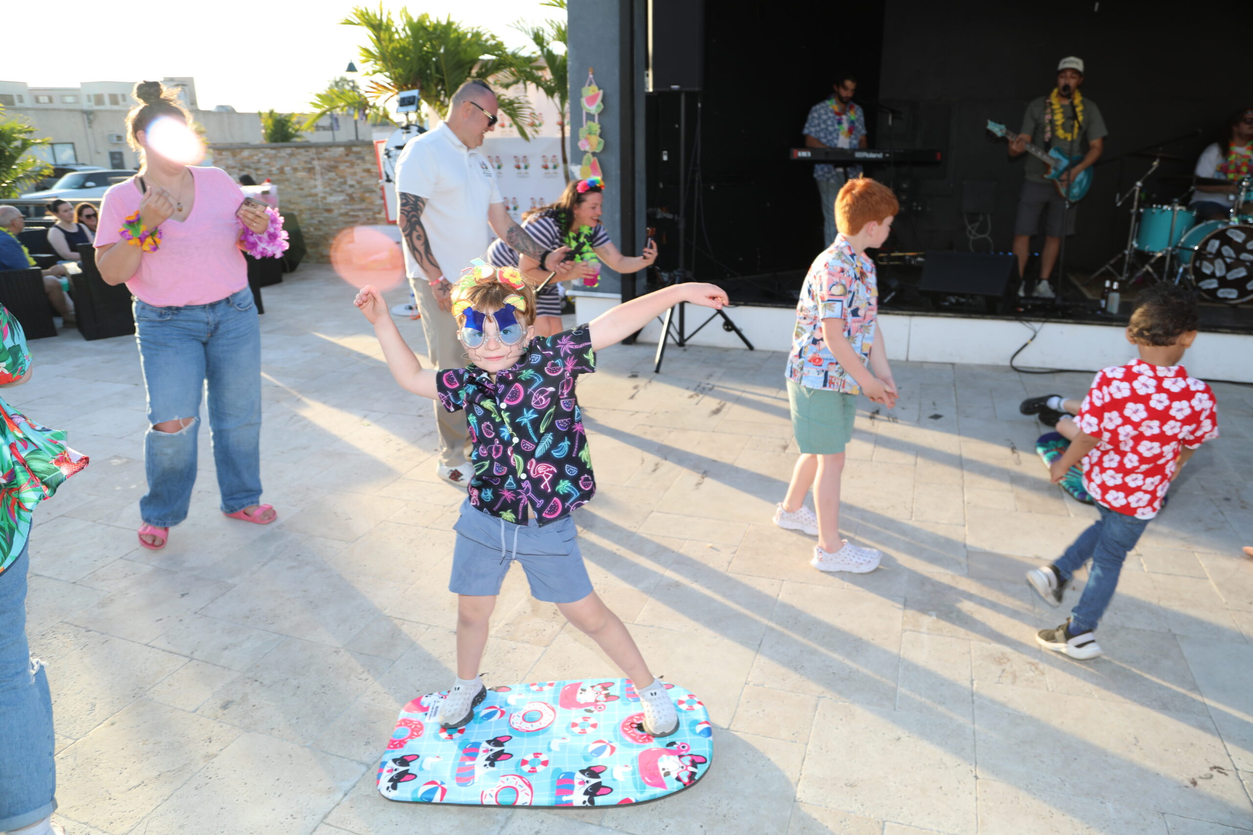 Children dancing at an outdoor party; a girl in a colorful tropical shirt and sunglasses stands on a patterned board as others move to the music with a live band playing on a stage in the background.