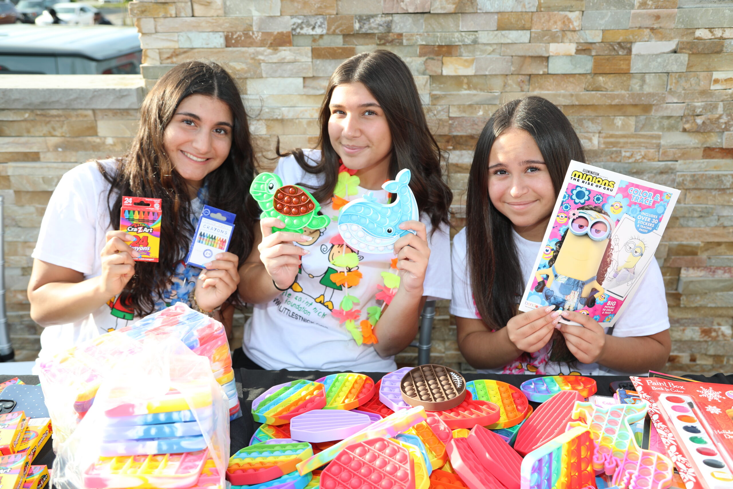 Three smiling girls pose at an outdoor table full of colorful toy kits and coloring books, showing their purchases against a stone wall backdrop.