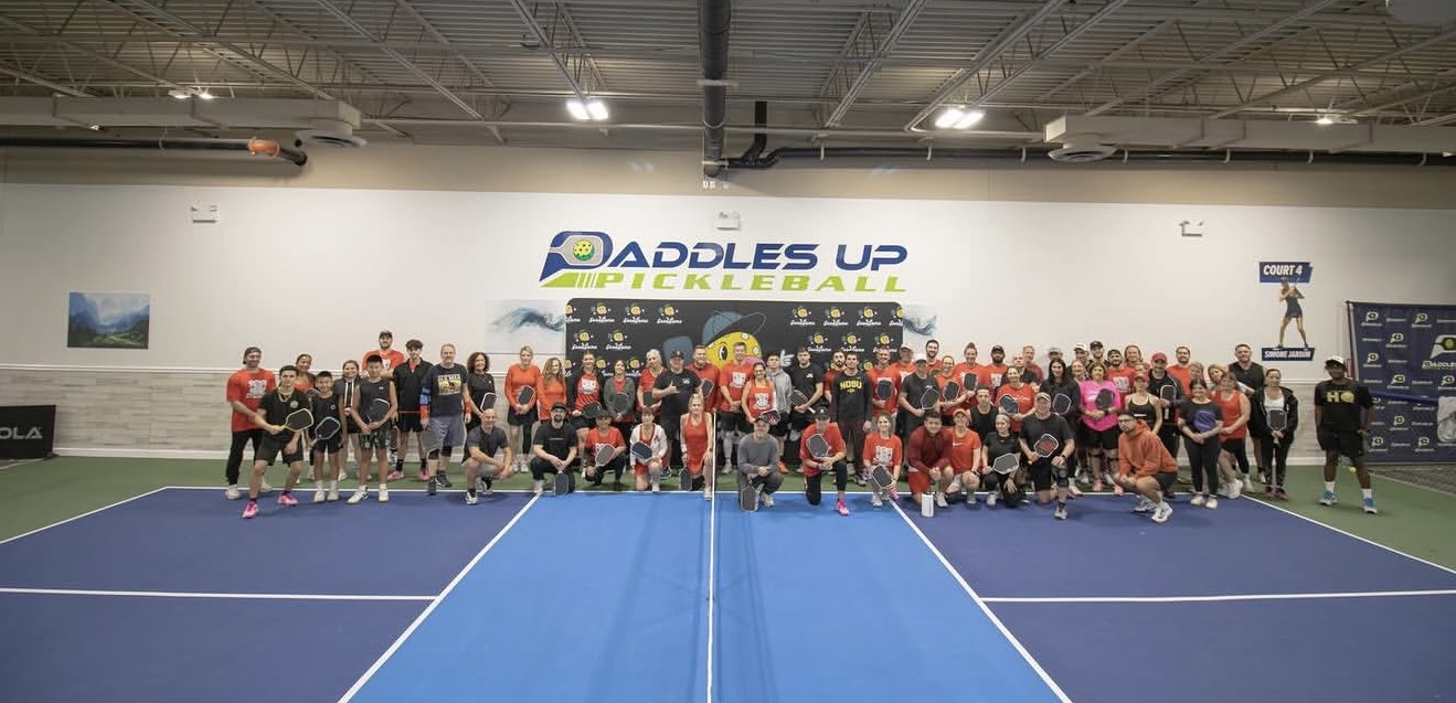 Group of pickleball players and coaches on an indoor court posing for a group photo in front of a banner reading 'Addles Up Pickleball'