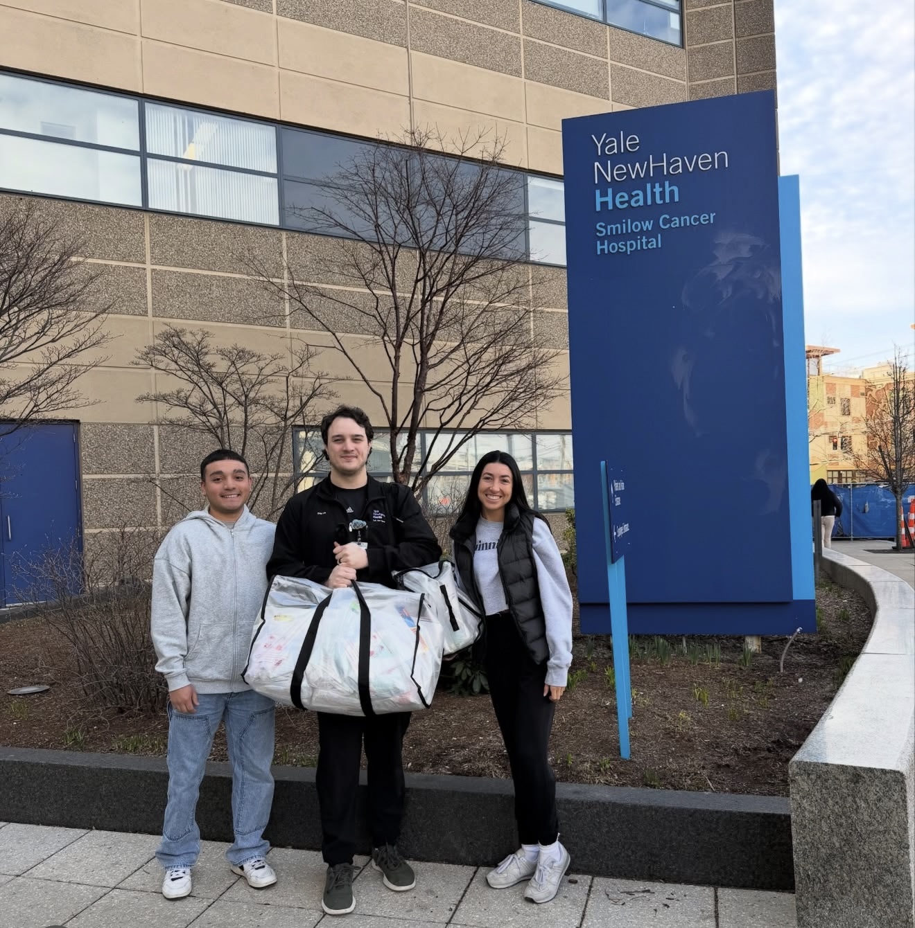 Three people pose outside Yale New Haven Health Smilow Cancer Hospital, smiling, with a large white duffel bag in the center near a tall blue sign.