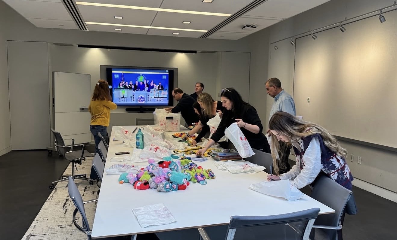 People stand around a long table, crafting and bagging plush toys in a conference room.