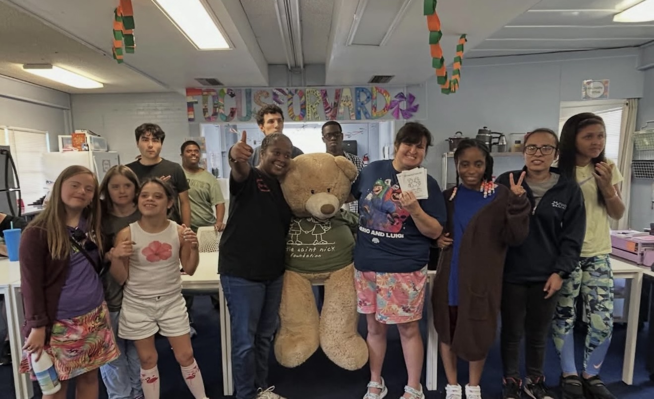 Group of diverse children and adults posing with a large teddy bear mascot in a bright classroom, some making peace signs, colorful banner in back