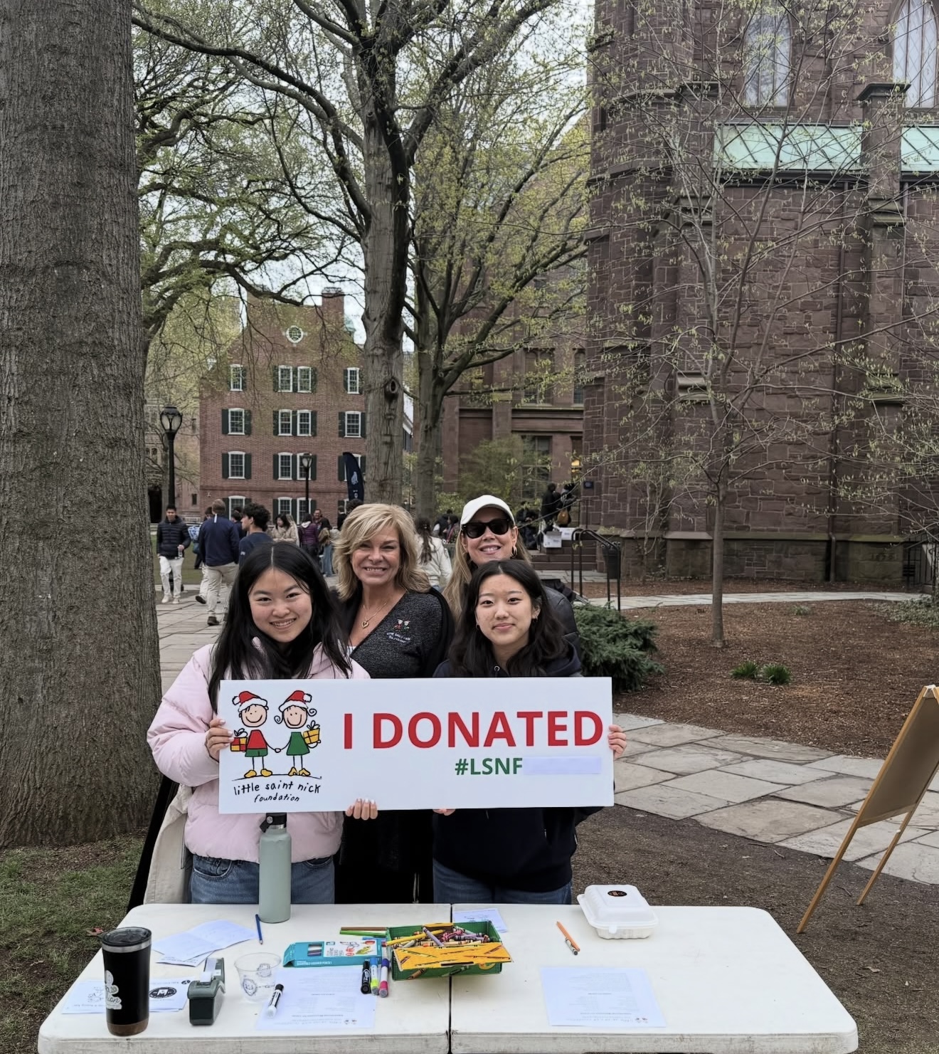 Group of smiling people holding a white sign that reads 'I DONATED' with '#LSNF' outdoors on a college campus; cartoon kids on sign; table with papers in foreground.