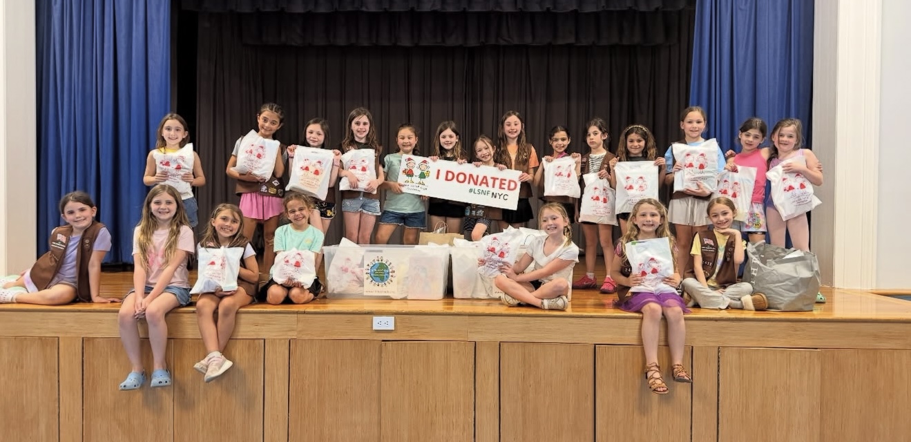 Group of smiling children on a stage holding donation bags and a banner that reads I DONATED (LSF NYC).