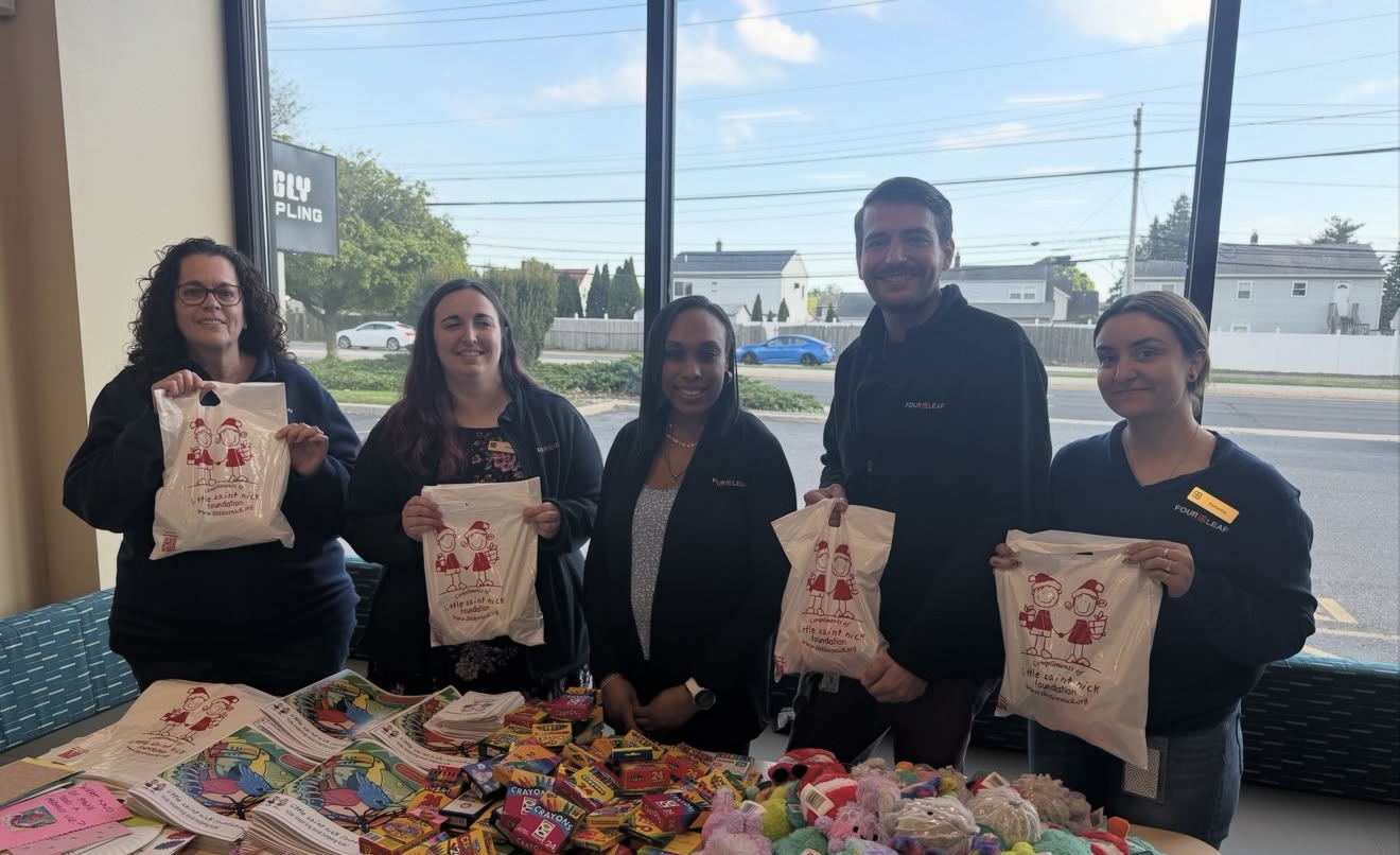 Five volunteers pose indoors by a large window, each holding a white tote bag with red cartoon figures; a table in front is filled with colorful toys and supplies.