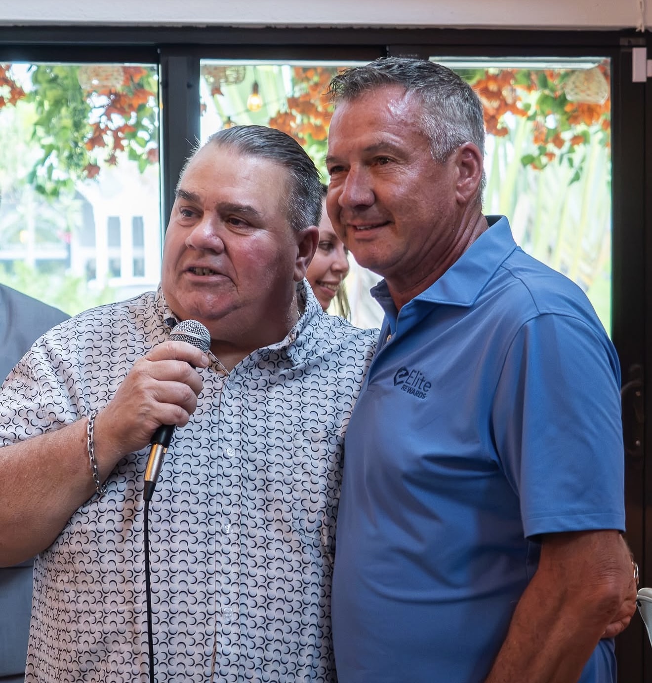 Two men at a social event: one holding a microphone and speaking, the other standing beside him in a blue polo with a logo visible.
