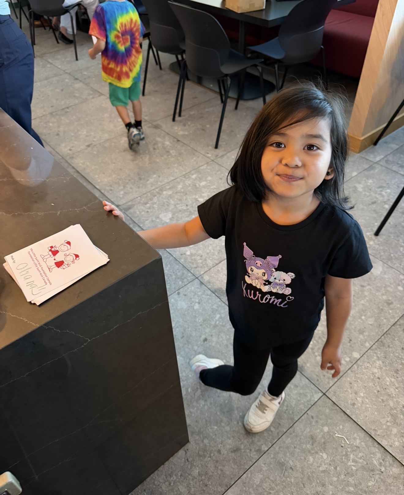 Young girl wearing a black Kuromi shirt stands at a dark counter, hand on the edge, smiling at the camera; a small white paper with red cartoon is on the counter.