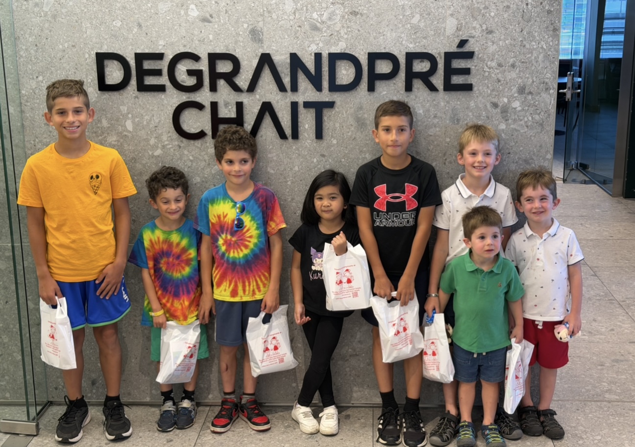 Seven children pose indoors in front of a grey wall with large black letters, each holding a white bag.