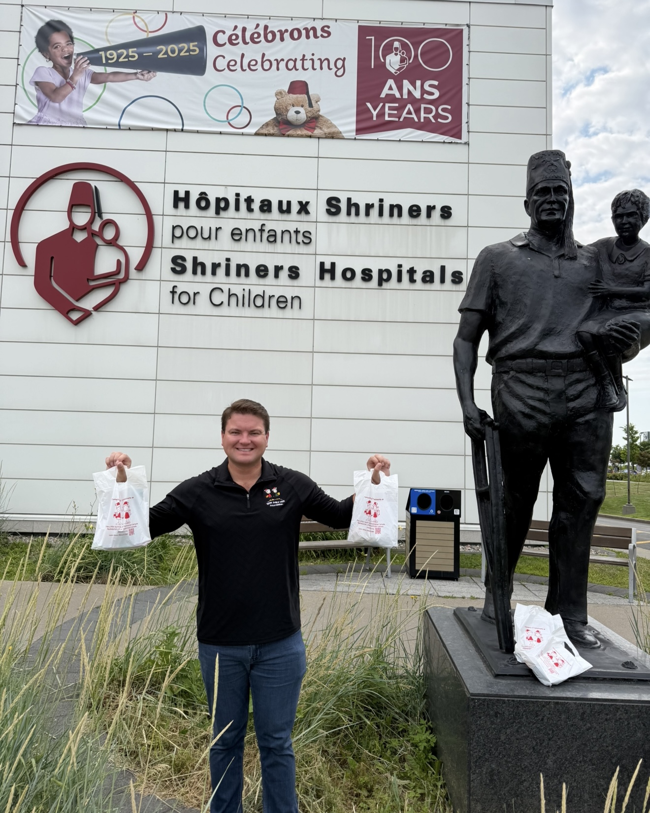 Man standing outside Shriner Hospitals for Children, holding two white gift bags, with a dark statue of a man and child nearby and a banner above.