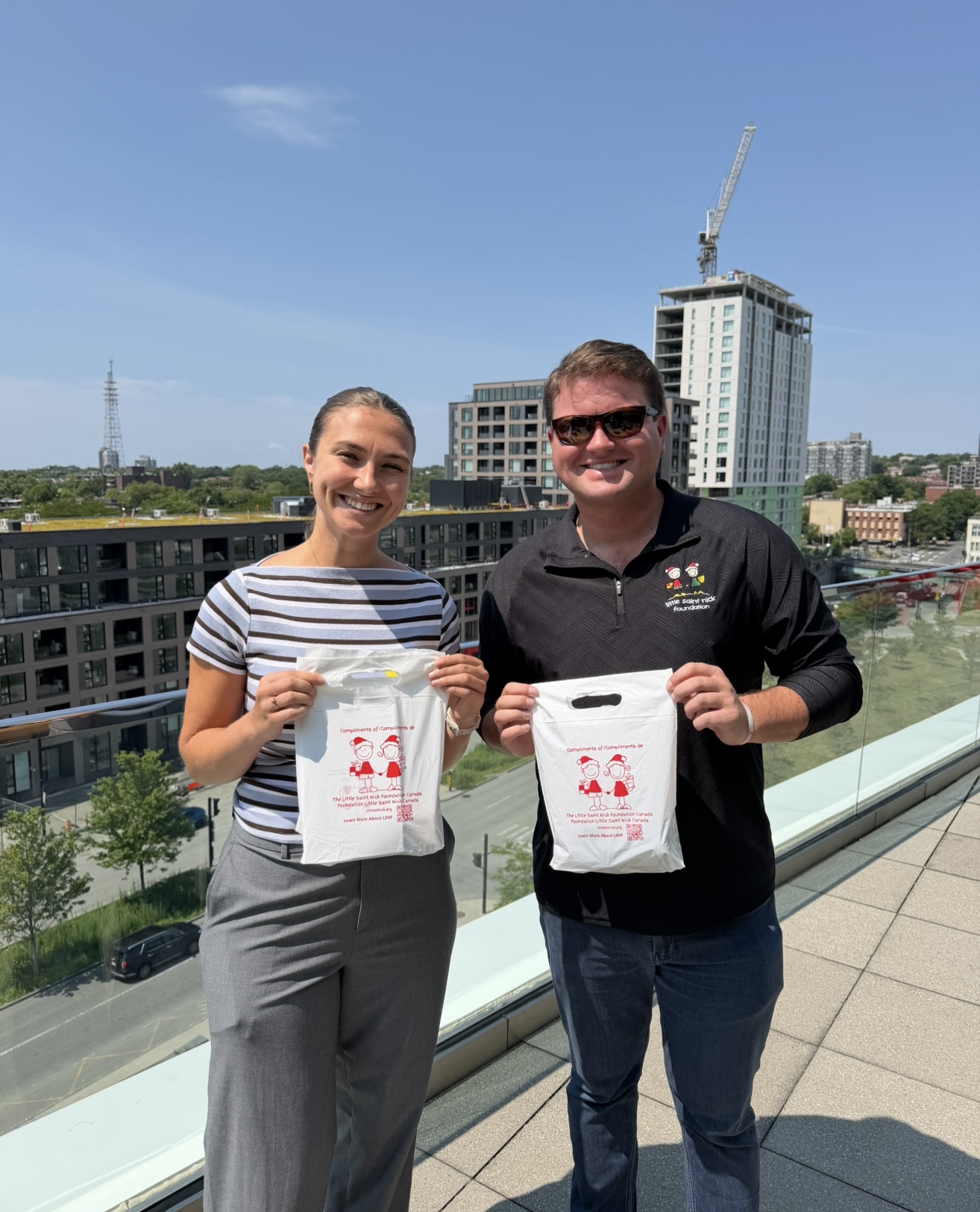 Two adults stand on a rooftop balcony, each holding a white bag with red print, smiling at the camera with a city skyline and construction crane behind them.