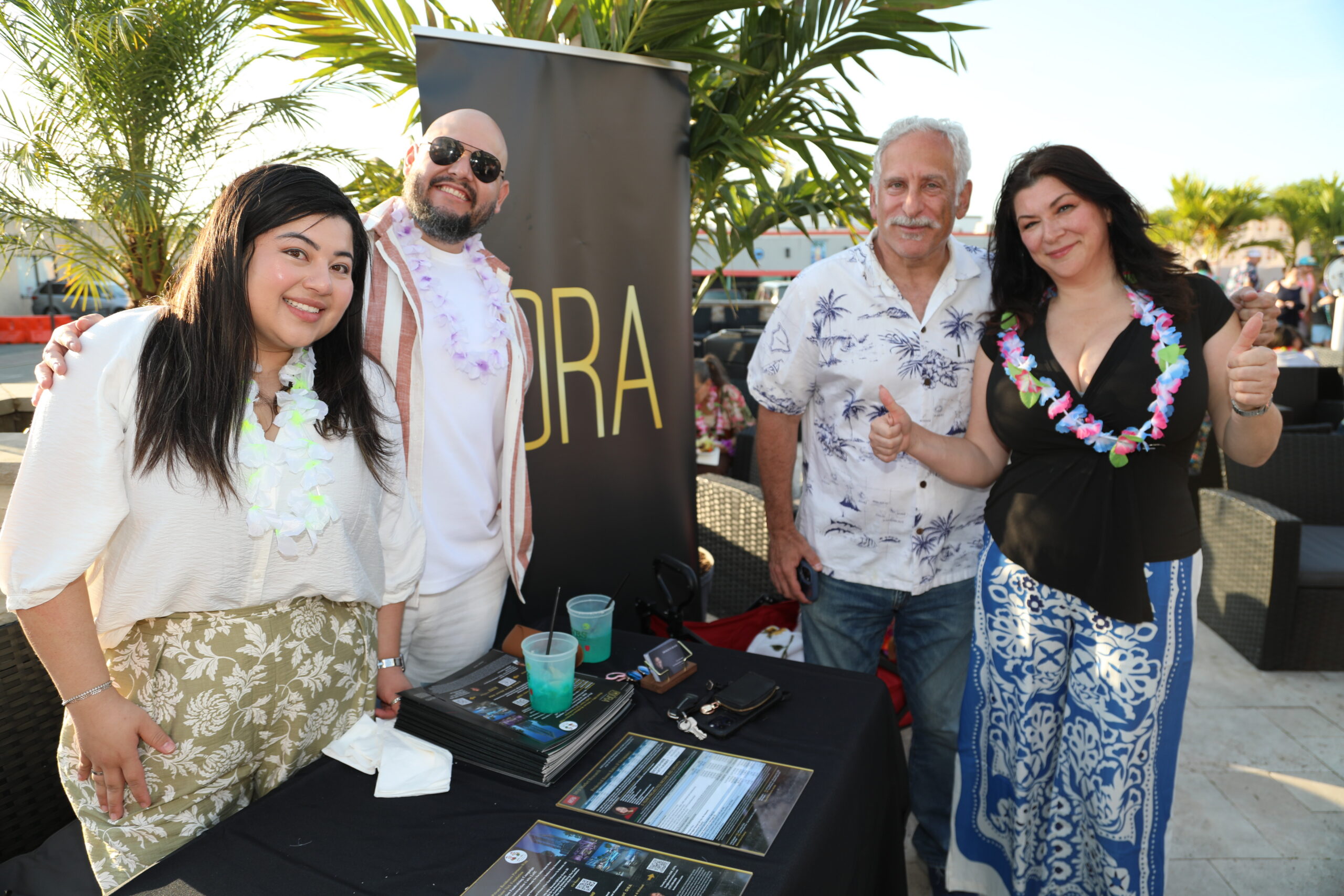 Four adults pose at an outdoor event booth, wearing Hawaii-style leis and casual summer clothing with brochures on a table in front of them.