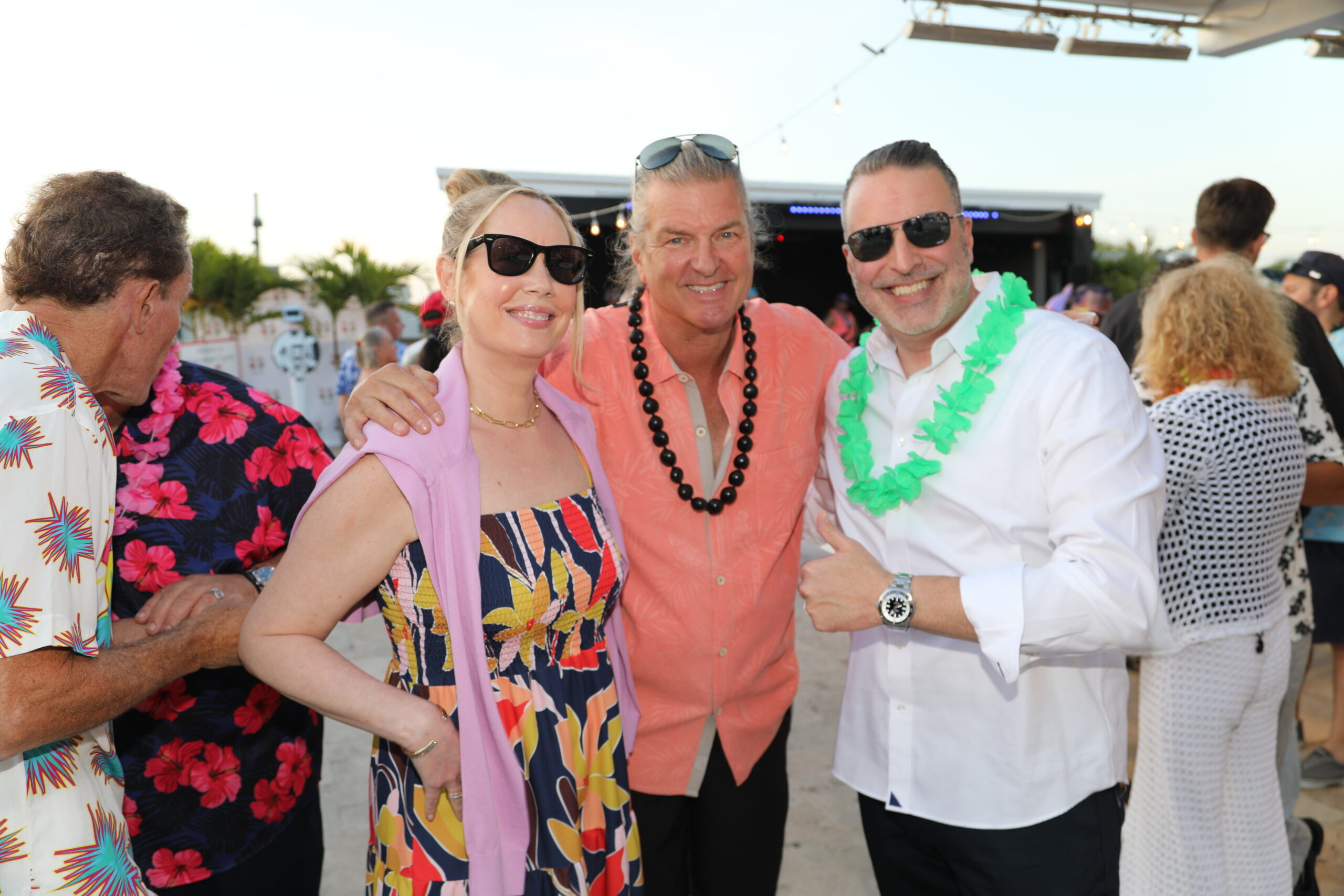 Group of friends at a tropical party posing for a photo, wearing colorful outfits and leis, smiling at the camera.
