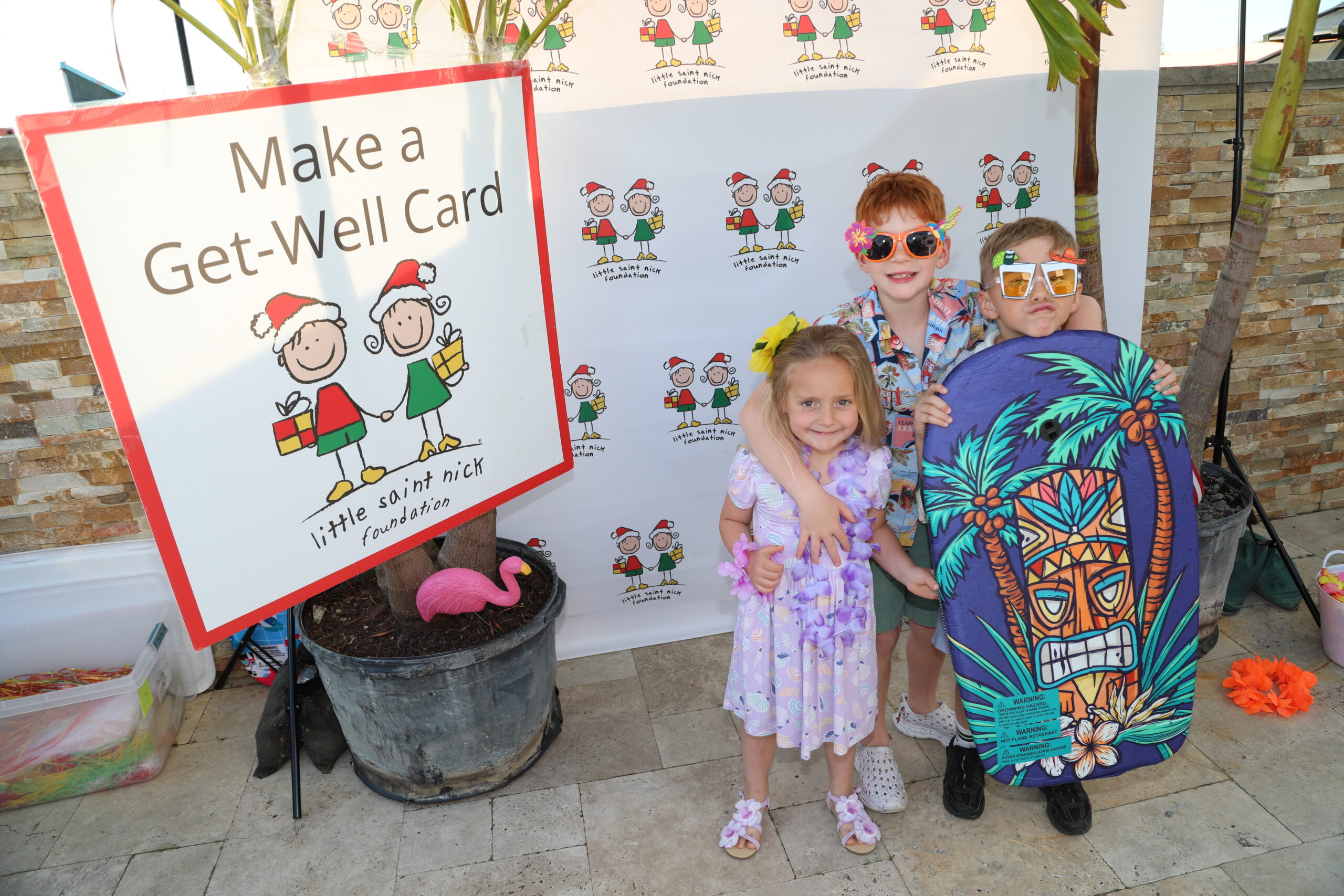 Children pose with a large 'Make a Get-Well Card' sign at a charity event, backdrop covered in festive logos; one girl wears a floral lei and pink dress.