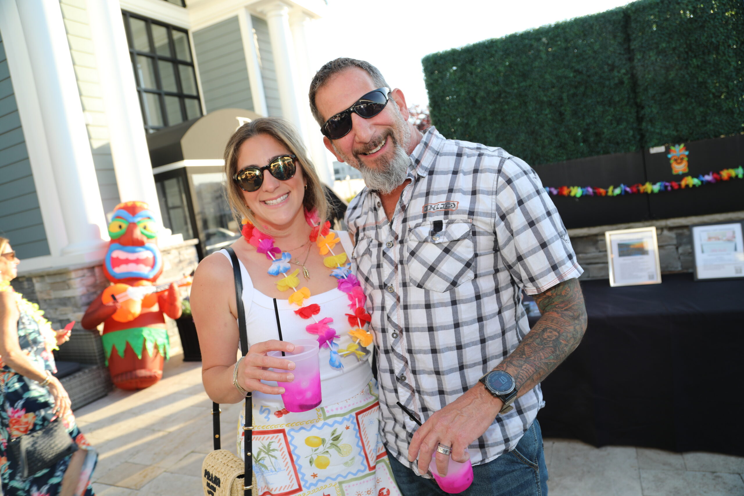 Smiling couple wearing sunglasses with leis, posing outdoors holding a pink drink; tiki figure and hedge in the background.