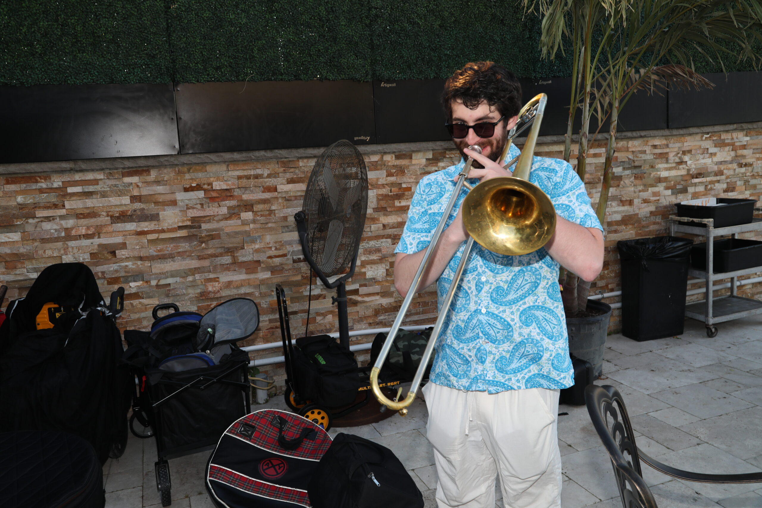 Man wearing a blue paisley shirt and sunglasses plays a trombone outdoors, surrounded by bags and equipment against a brick wall.