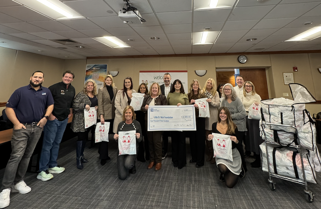 Group of adults posing in a meeting room holding a large donation check and white gift bags, smiling proudly.