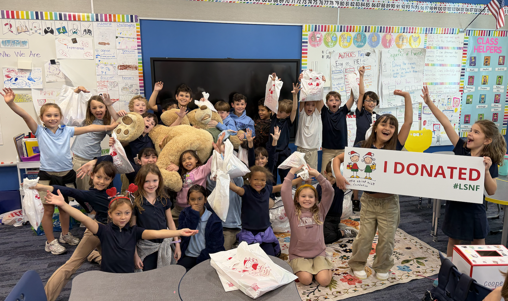 Cheerful group of elementary students in a classroom raising plastic bags of donations; a large teddy bear sits center, and two kids hold a sign reading 'I DONATED' with '#LSNF' on the right.