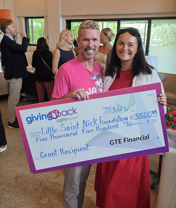 Smiling man in a pink shirt and woman in red dress hold a large ceremonial check from a charity event, posing for the photo with others in the background.