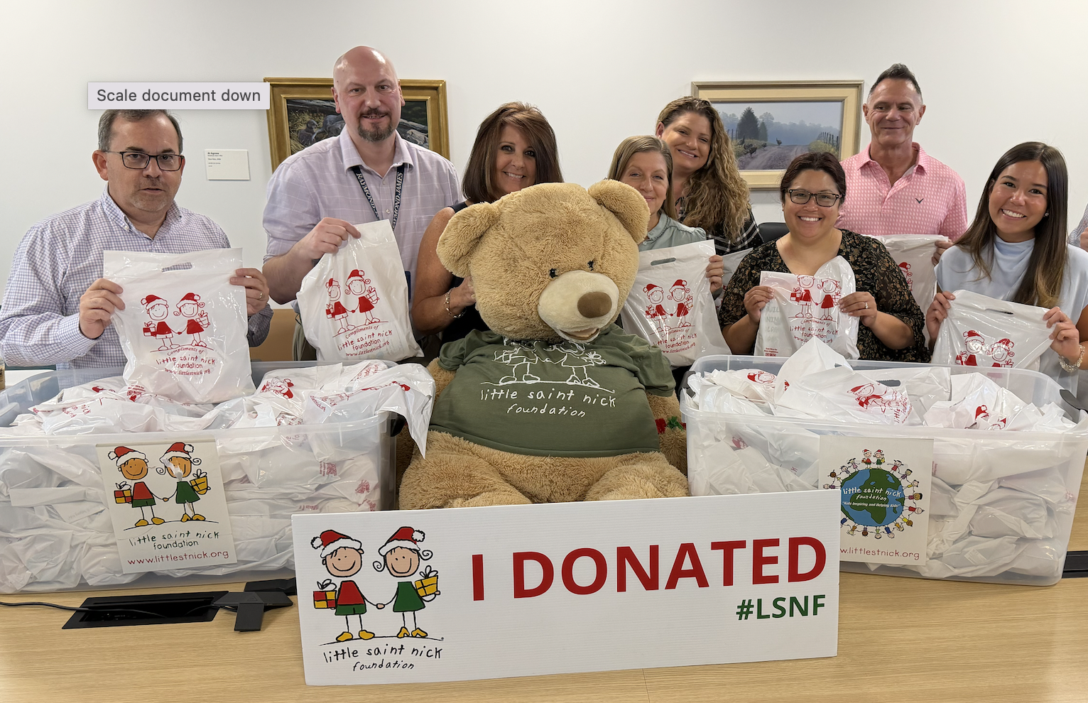 Group of people posing around a large teddy bear wearing a Little Saint Nick Foundation shirt, holding donation bags.
