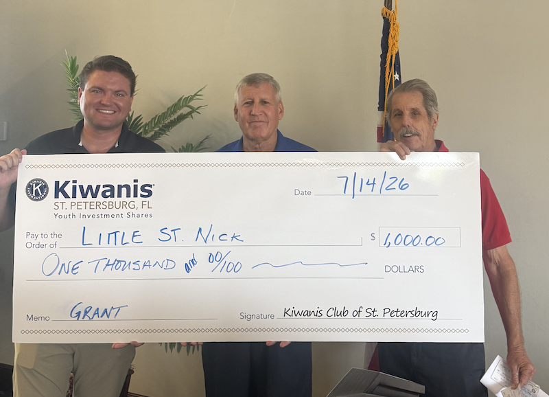 Three men stand holding a large Kiwanis charity check made out to Little St. Nick for $1,000, dated 7/14/26, at a club presentation.