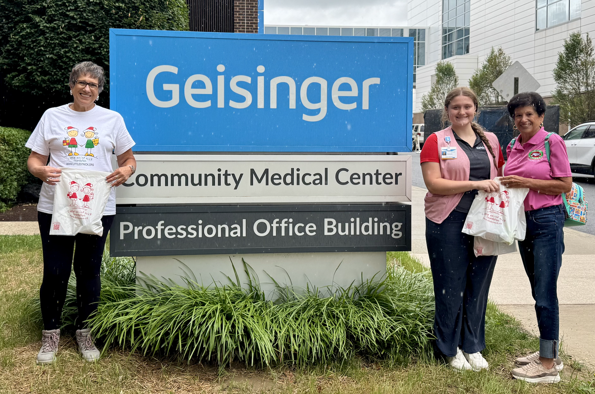 Three women stand outdoors beside a Geisinger Community Medical Center sign, each holding a white tote bag.