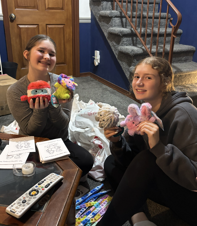 Two smiling girls sit on the floor holding colorful stuffed animals, with crafts and a remote on a nearby table in a cozy room.